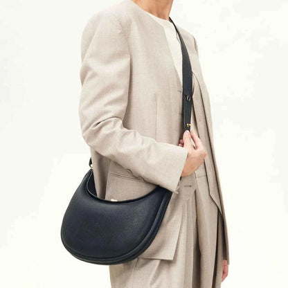 Person wearing a beige coat holding a black crossbody handbag on a white background